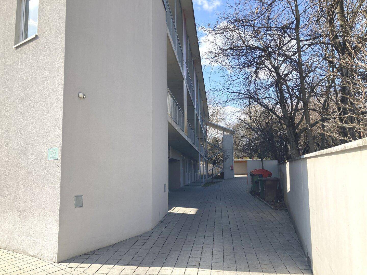 Lange Fassade eines modernen Gebäudes mit Balkonen und gepflastertem Zugangsbereich bei Sonnenschein.
