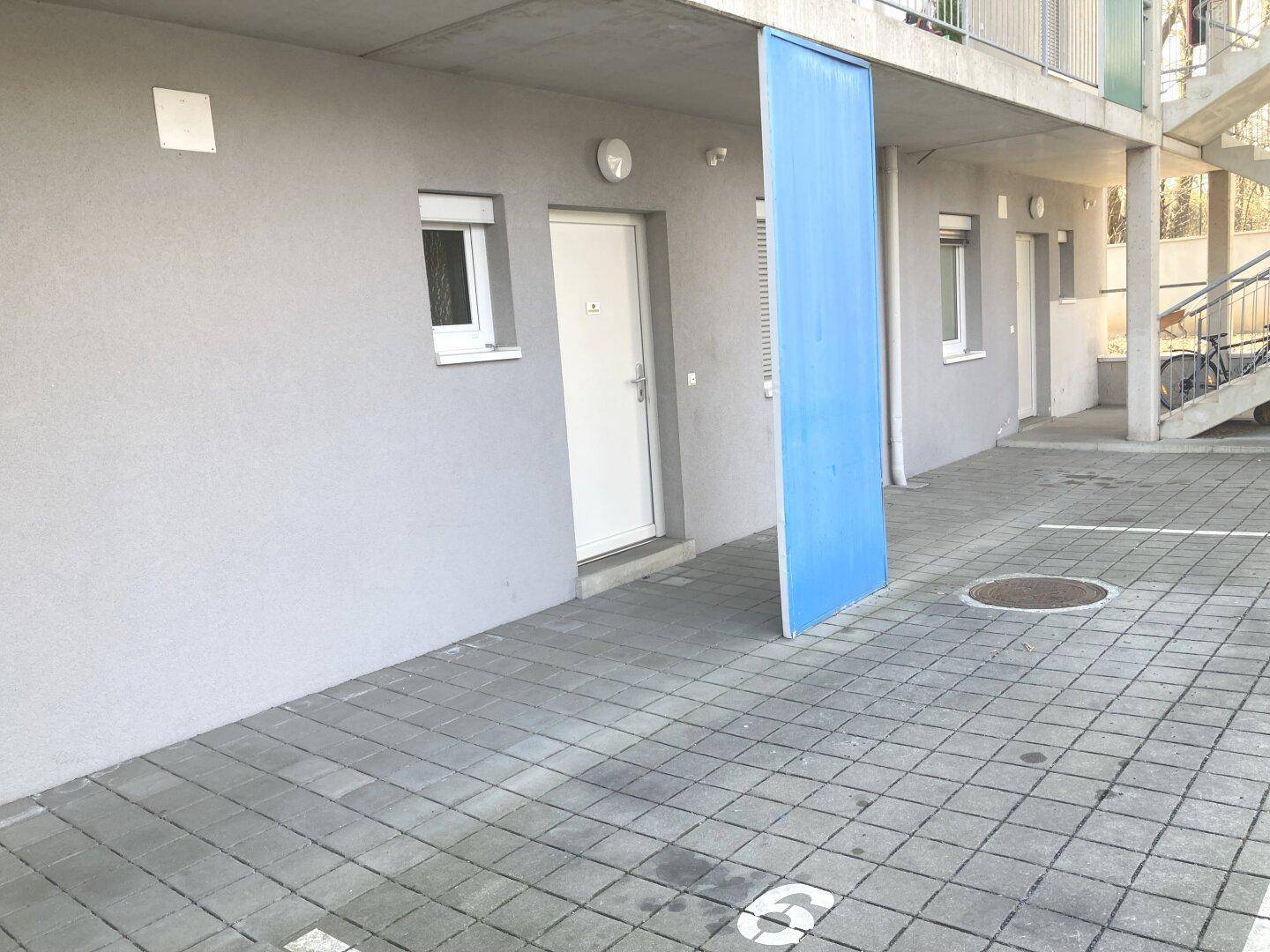 Eingangsbereich eines modernen Gebäudes mit gepflastertem Vorplatz und Parkplätzen.