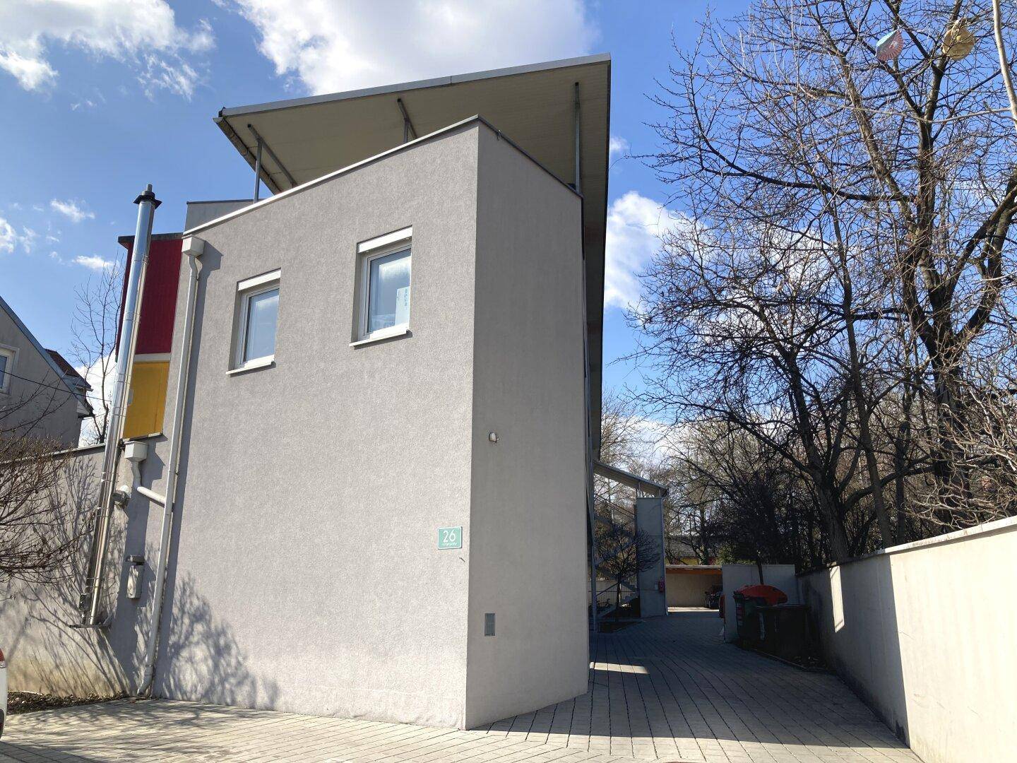Modernes Gebäude mit grauer Fassade, Flachdach und Bäumen im Hintergrund.