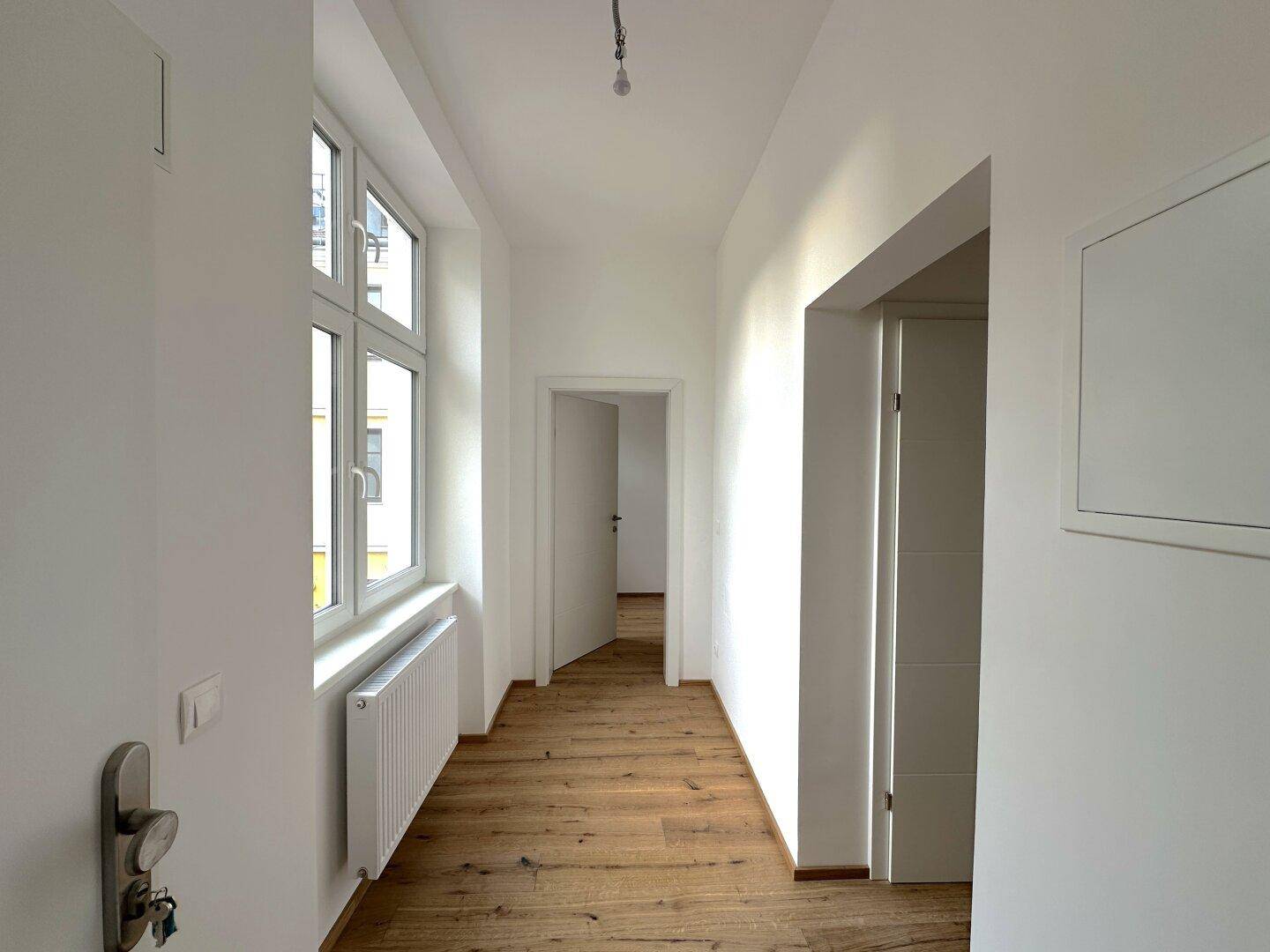 Langer, heller Flur mit Holzboden, großem Fenster und Heizkörper, der zu weiteren Räumen führt.