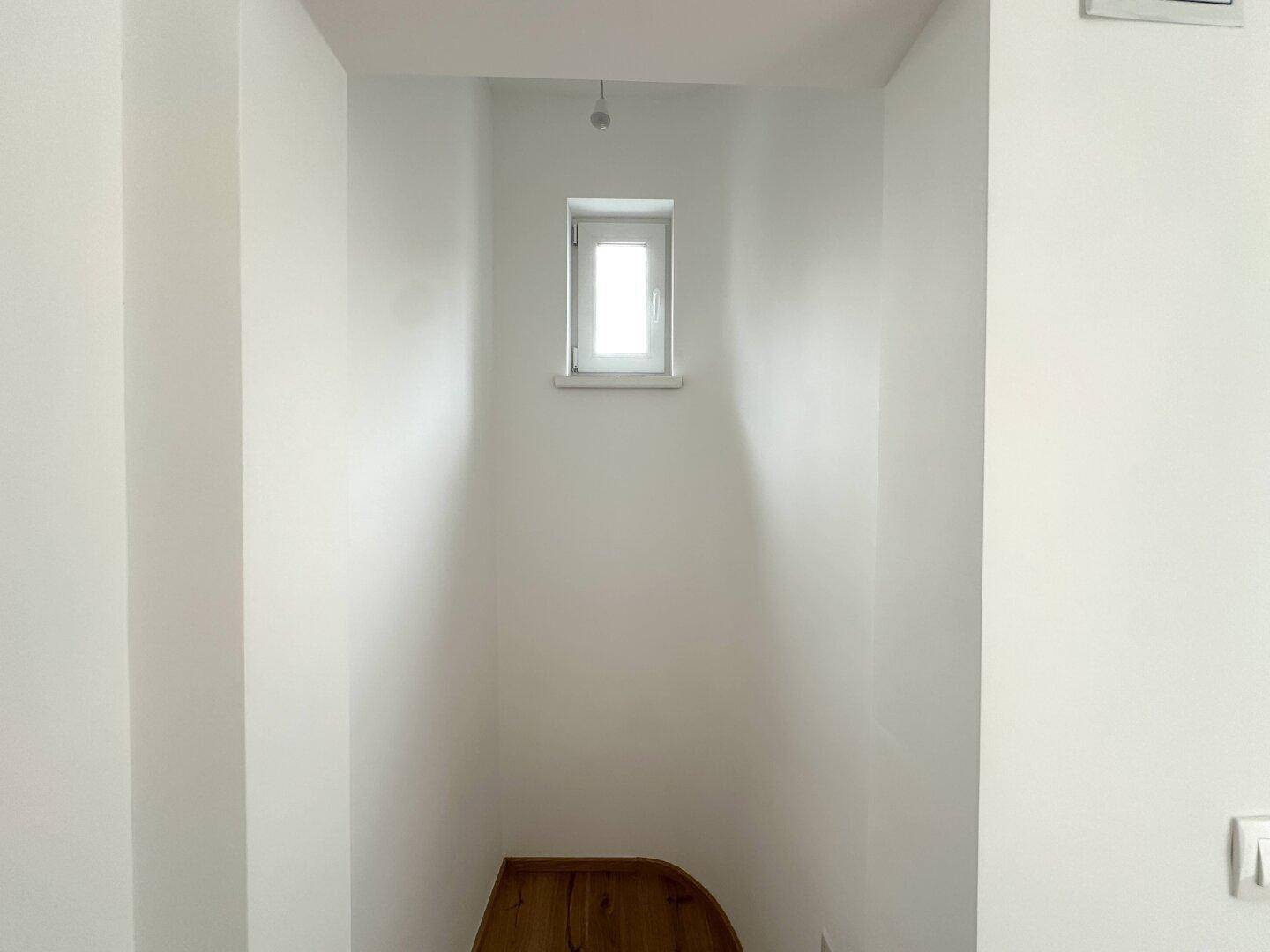 Schmaler, heller Bereich mit einem kleinen Fenster und einer Holzstufe, möglicherweise ein Treppenabsatz.