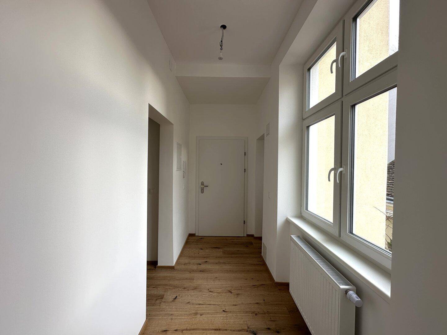 Eingangsbereich mit Holzboden, großem Fenster, Heizkörper und der Wohnungstür, die viel Licht hereinlässt.
