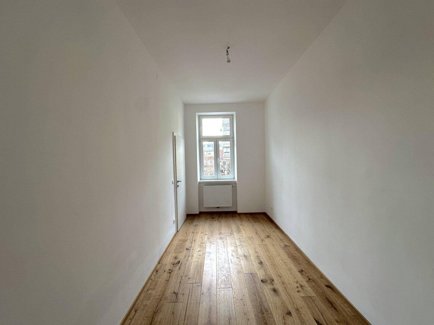Helles, leeres Zimmer mit Holzboden und einem Fenster, das viel Tageslicht hereinlässt.