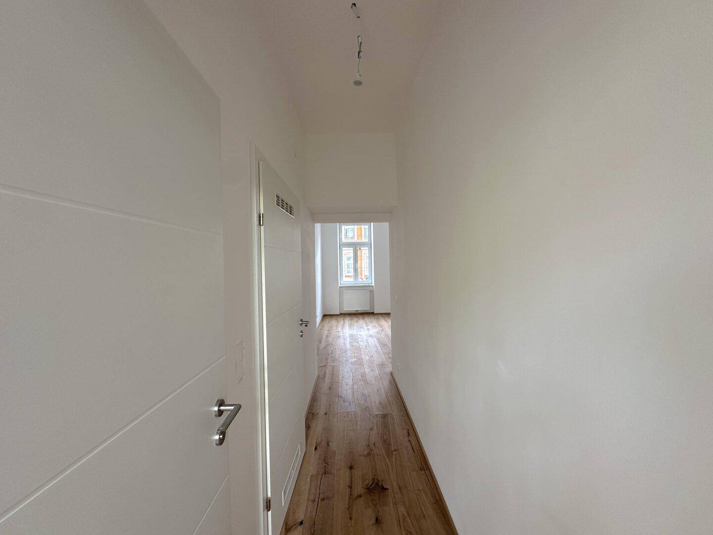 Langer Flur mit Holzboden und weißen Wänden, der zu einem hellen Zimmer am Ende führt.