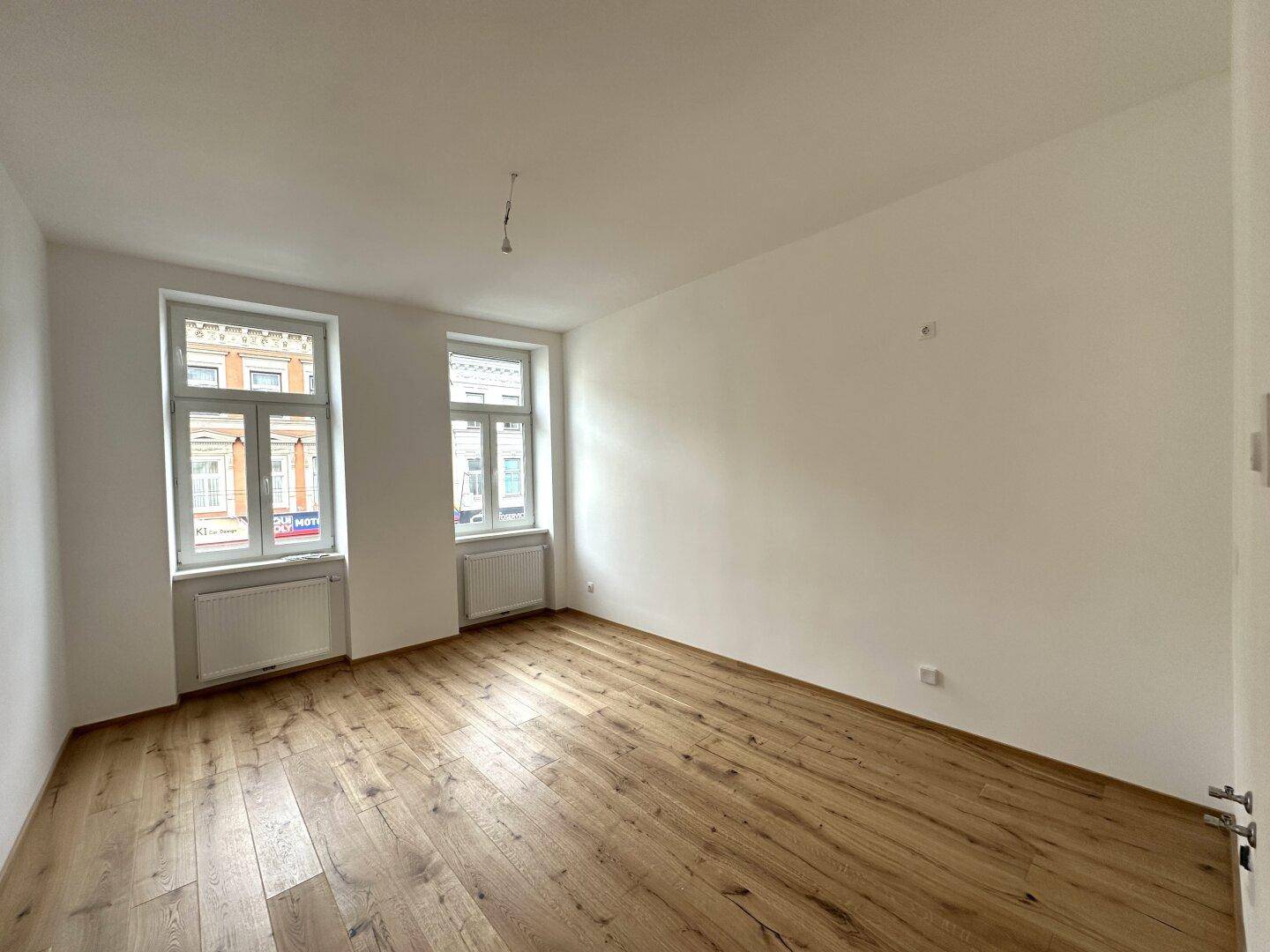 Heller, leerer Raum mit Holzboden und zwei großen Fenstern, die viel Licht spenden.