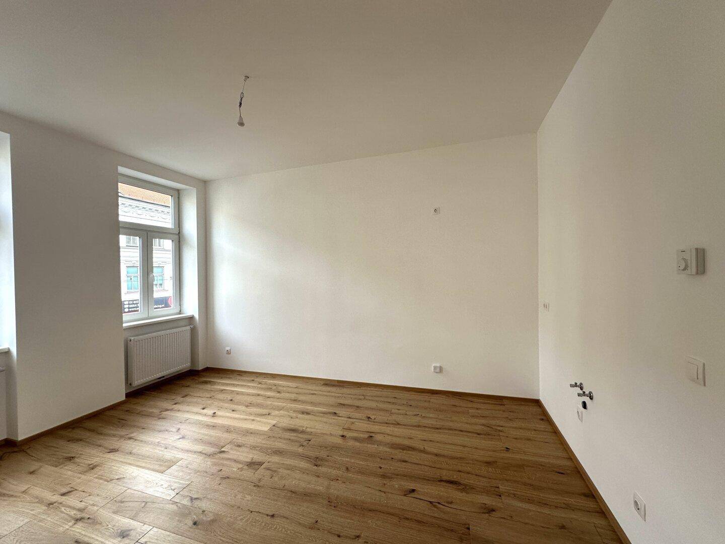 Leerer Raum mit Holzboden und Fenster, vorbereitet für eine Küche mit sichtbaren Anschlüssen.