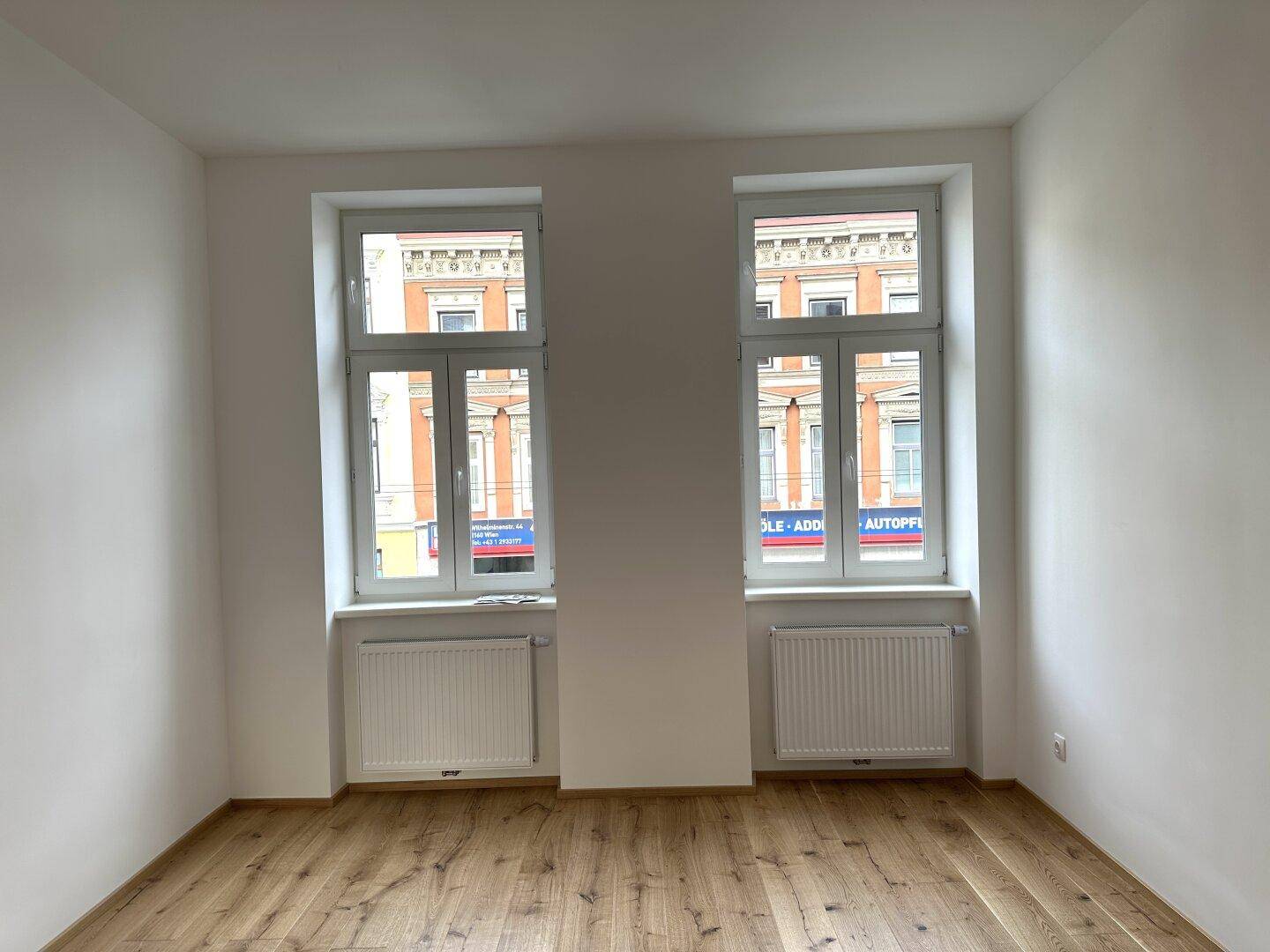 Geräumiger, heller Raum mit Holzboden und zwei Fenstern, die einen Blick nach draußen ermöglichen.
