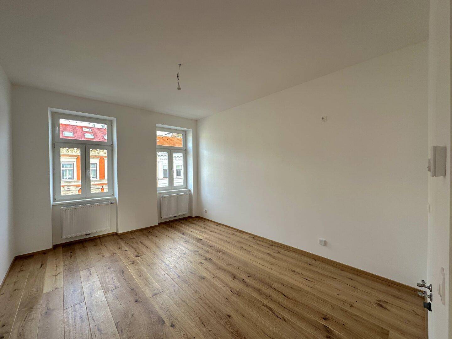 Geräumiges, helles Zimmer mit Holzboden und zwei Fenstern, die einen Blick auf die umliegenden Gebäude bieten.