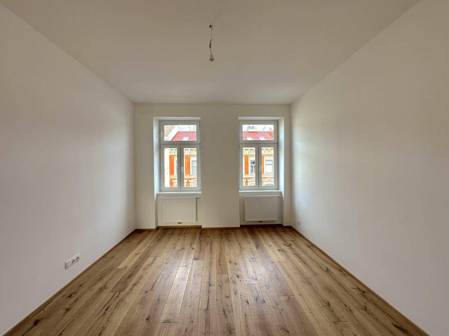 Helles, leeres Zimmer mit neuem Holzboden und zwei Fenstern, die auf die Straße blicken.