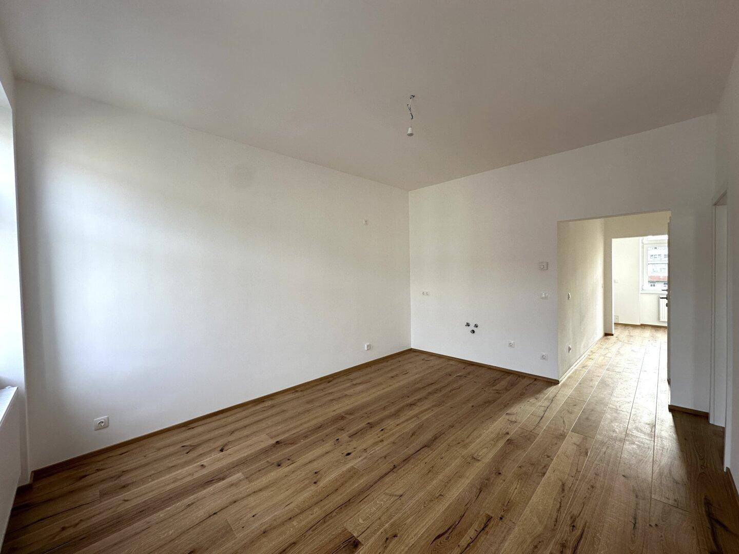Leeres Zimmer mit Holzboden und weiß gestrichenen Wänden, mit Blick auf einen angrenzenden Raum.