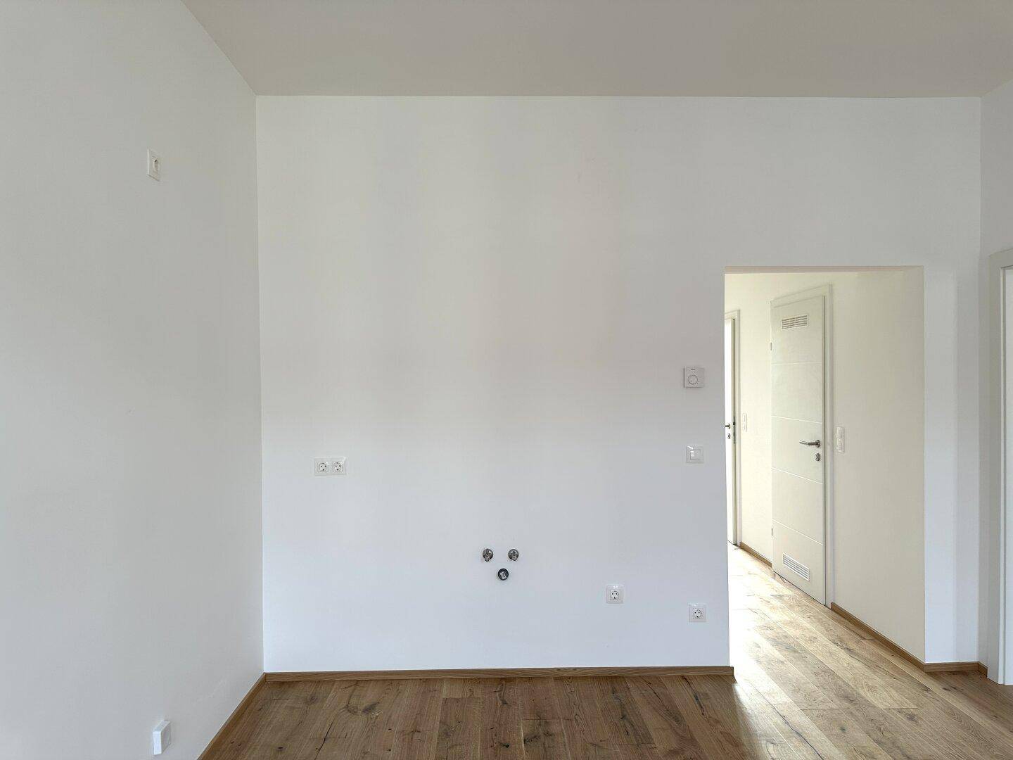Heller Raum mit Holzboden und weiß gestrichenen Wänden, mit Türzugang zu weiteren Zimmern.