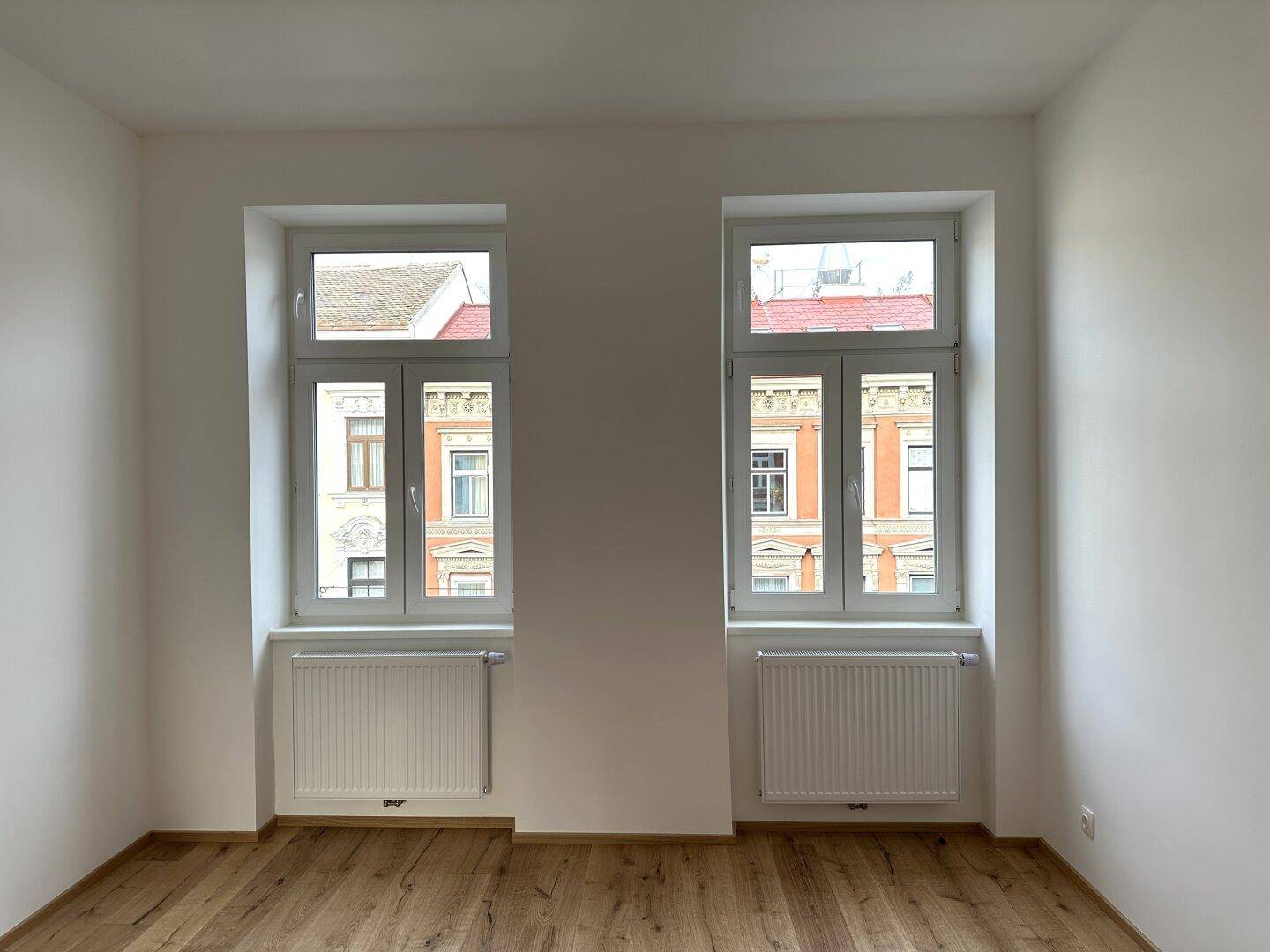 Zwei große Fenster in einem leeren Zimmer mit Holzboden, die viel Tageslicht hereinlassen.
