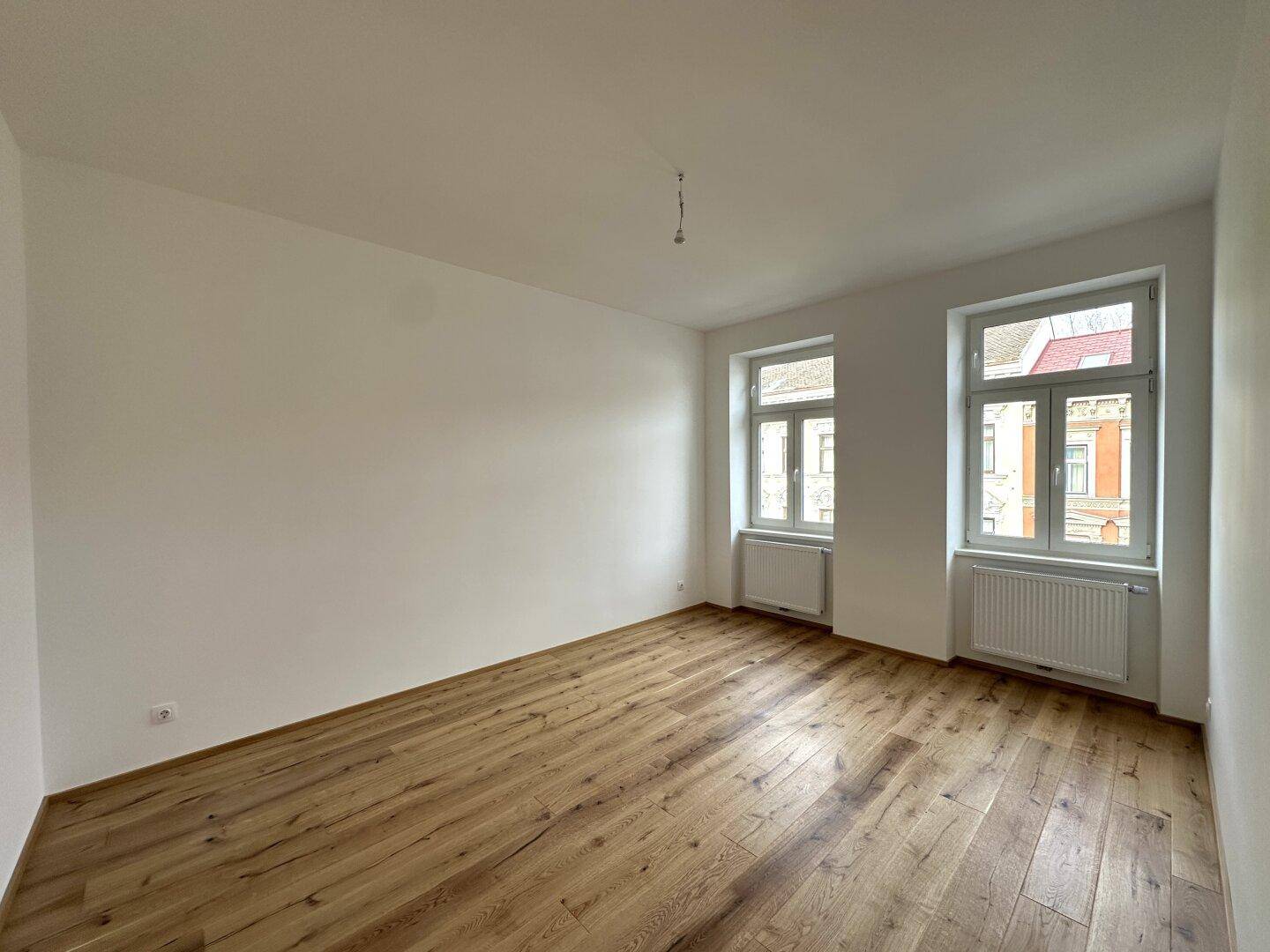 Geräumiges, helles Zimmer mit Holzboden und zwei Fenstern, die einen Blick auf die umliegenden Gebäude bieten.