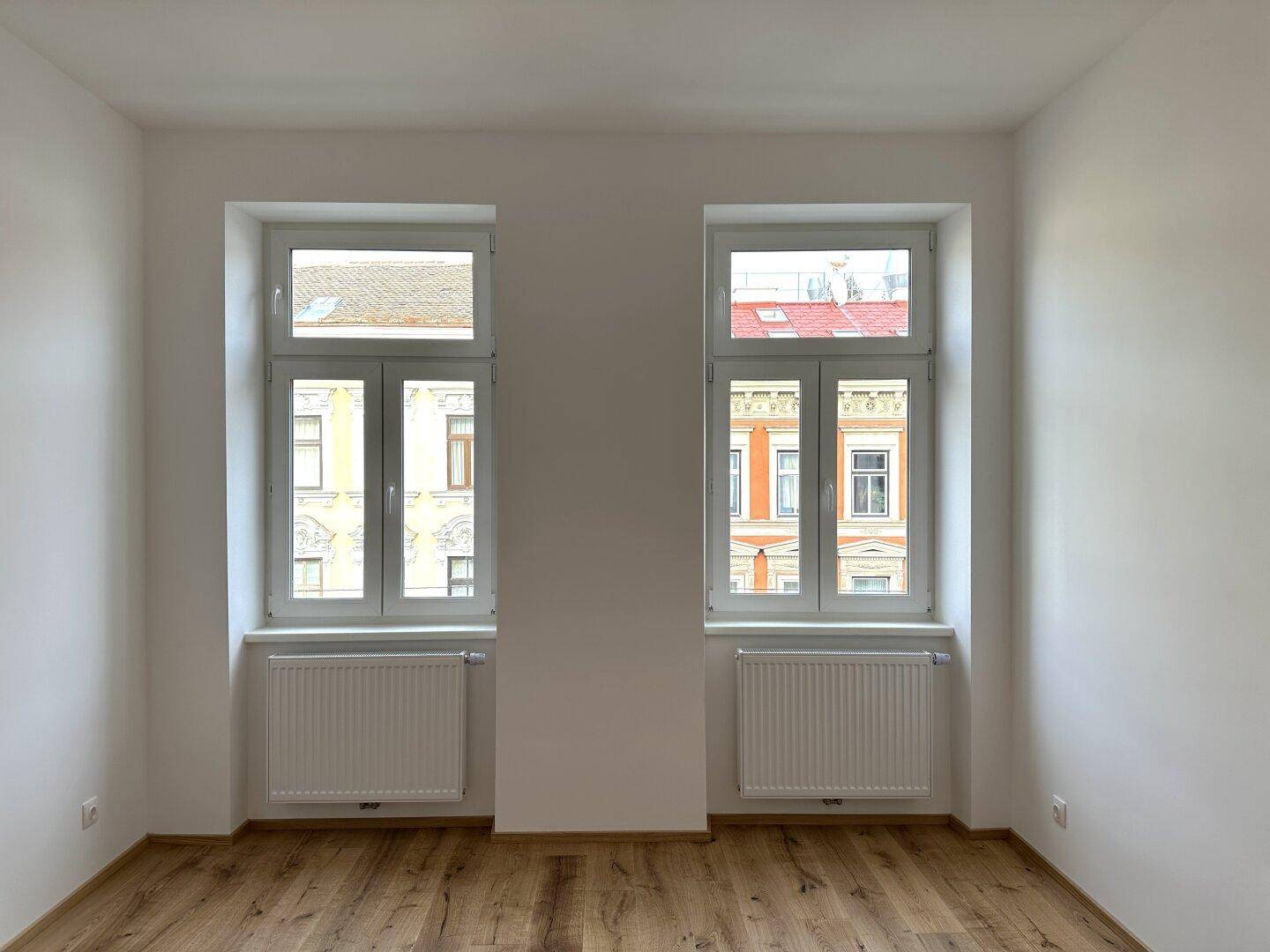 Zwei große Fenster in einem leeren Zimmer mit Holzboden, die viel Tageslicht hereinlassen.