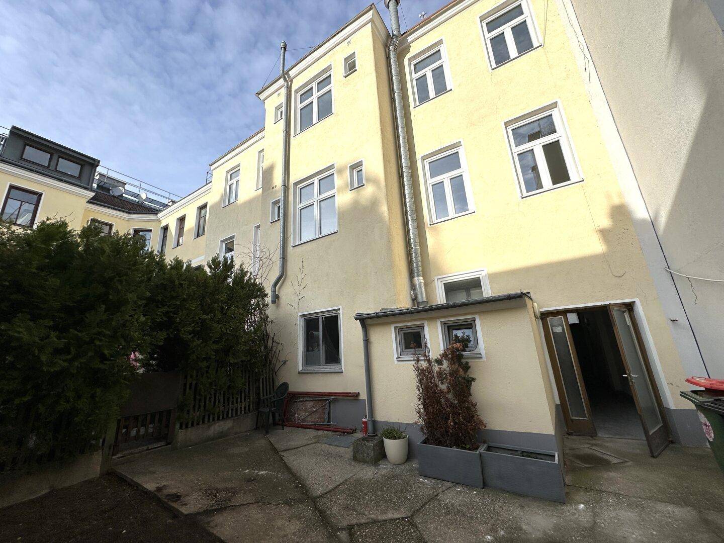 Hinterhofbereich eines gelben Gebäudes mit gepflastertem Boden, Sträuchern und Mülltonnen.