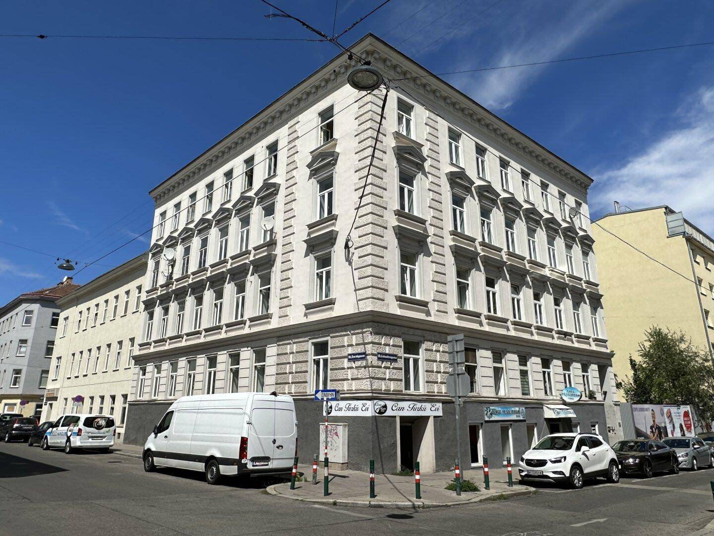 Eckgebäude mit klassischer Architektur und Geschäften im Erdgeschoss an einer städtischen Kreuzung unter blauem Himmel.