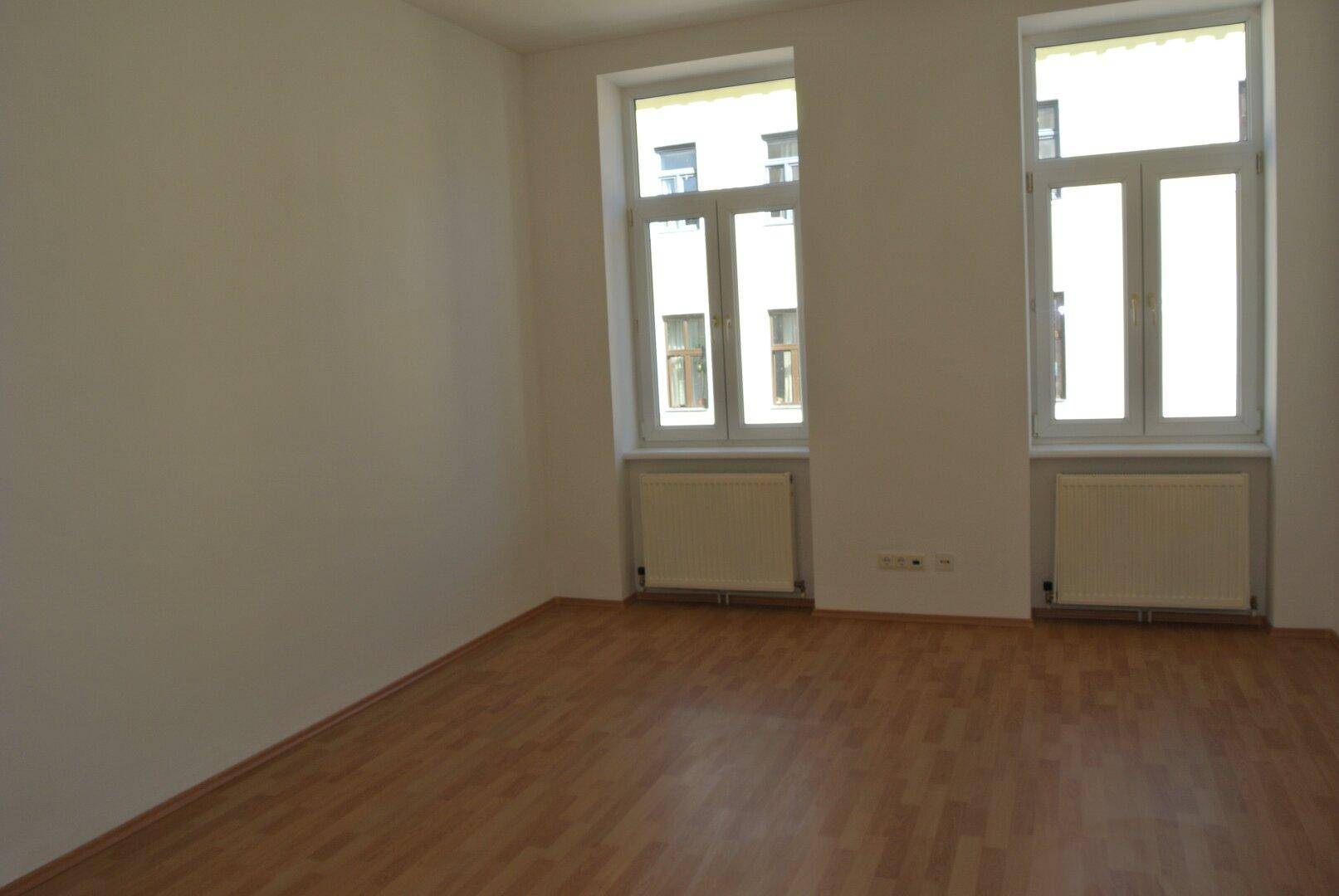 Leerer Raum mit zwei Fenstern, Heizkörpern und hellem Laminatboden, bereit zur Einrichtung.