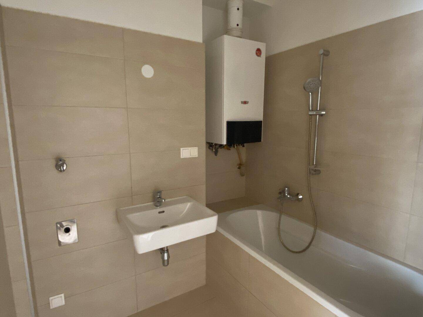 Badezimmer mit Badewanne, Waschbecken und Gastherme, gefliest in beigen Farbtönen.
