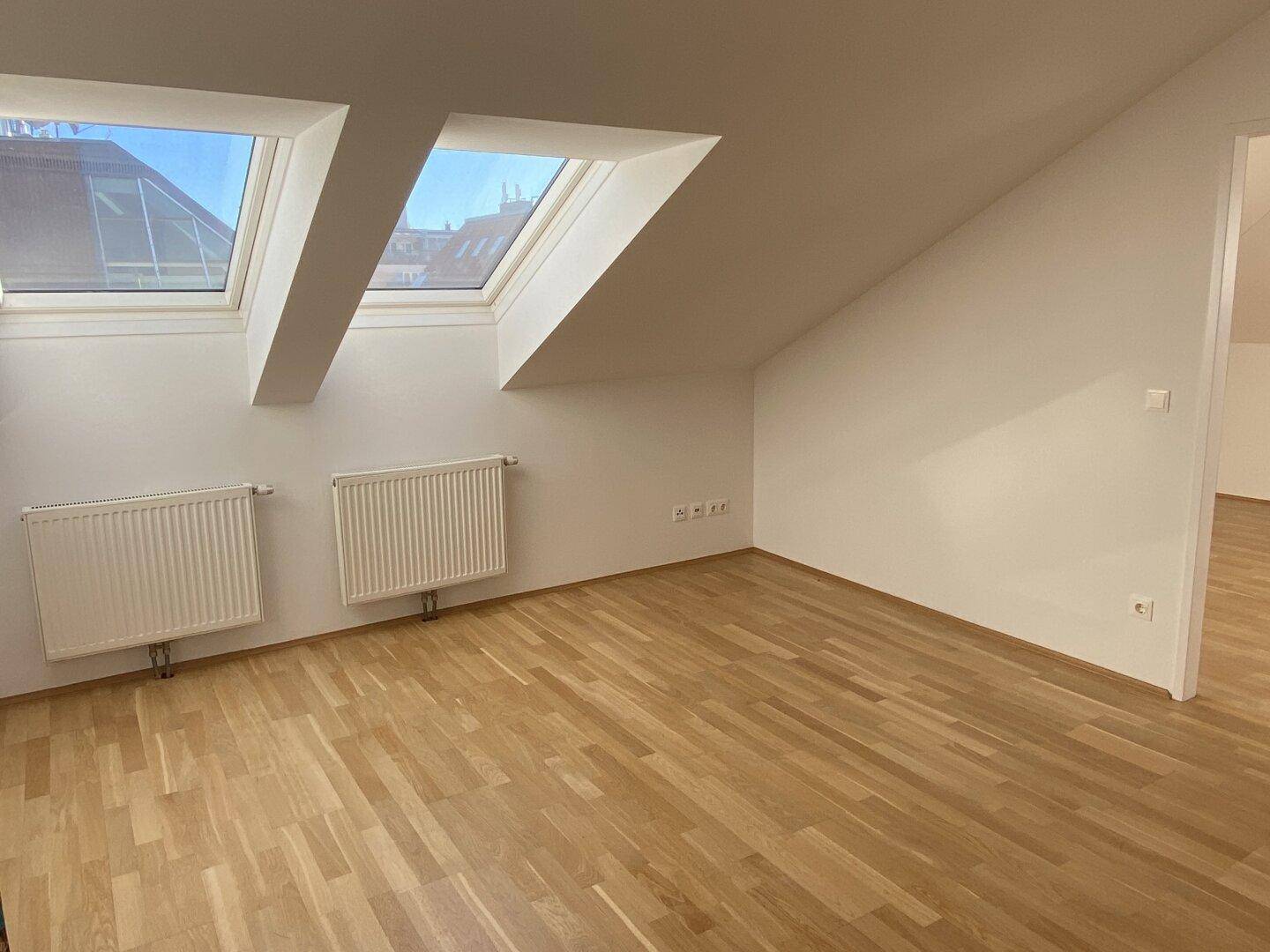 Heller Dachgeschossraum mit schrägen Wänden, zwei Dachfenstern und hellem Parkettboden.