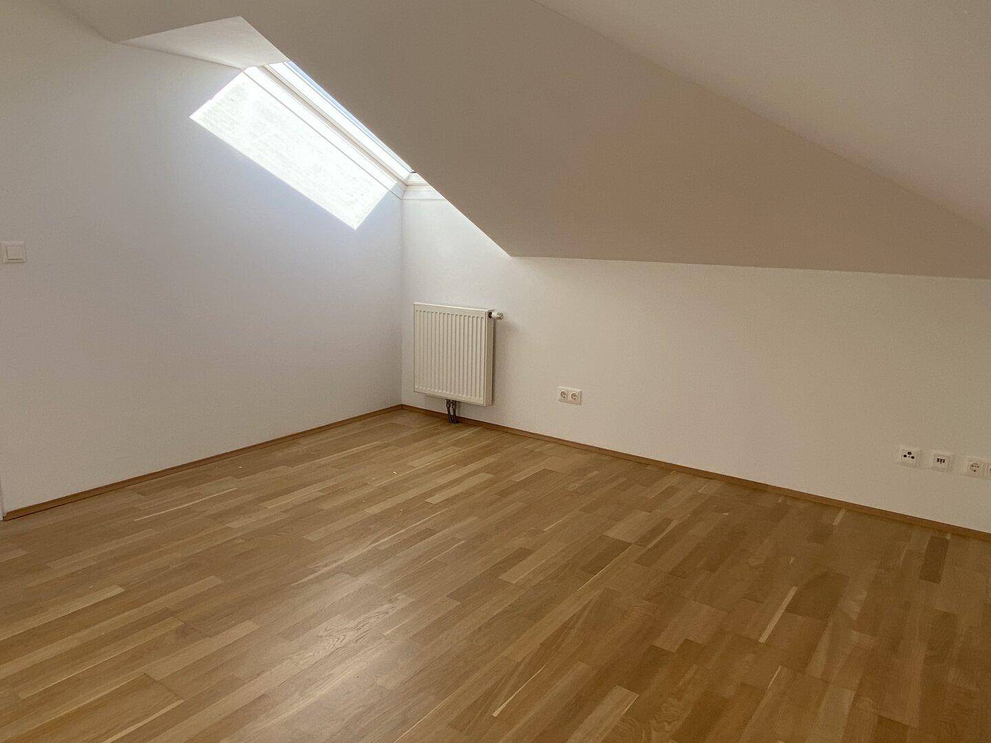 Dachgeschossraum mit schräger Decke, einem Dachfenster und hellem Holzparkettboden.