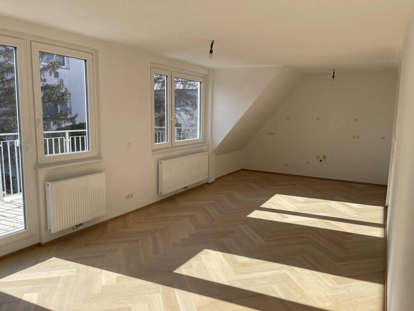 Heller, leerer Raum mit Fischgrätparkett, großen Fenstern und Zugang zum Balkon.
