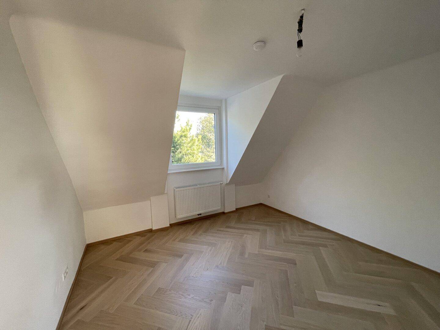 Gemütliches Zimmer mit Dachschräge, Fischgrätparkett und einem Fenster mit Grünblick.