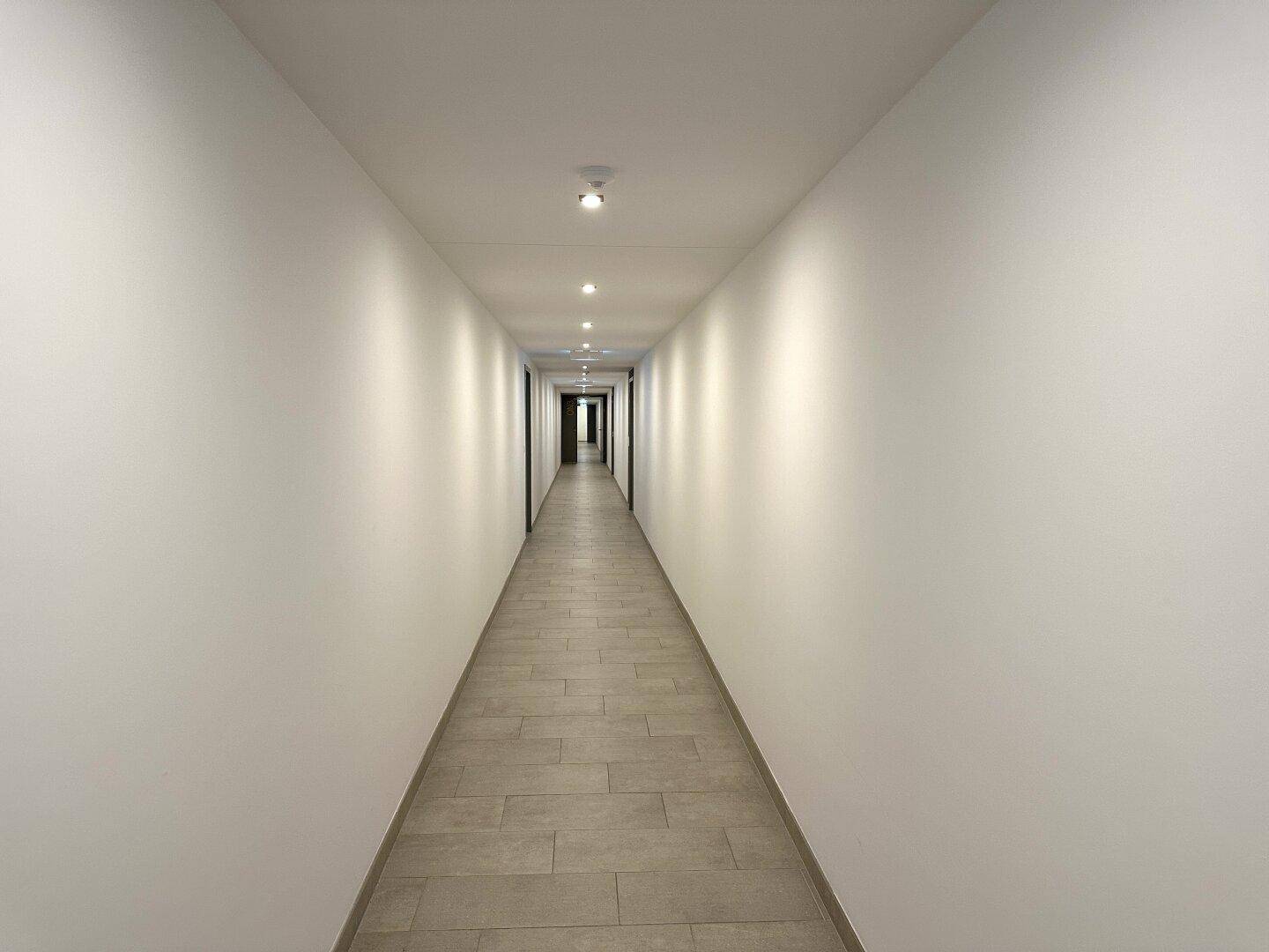 Langer, heller Flur mit Fliesenboden und modernen Deckenleuchten im Gebäude.