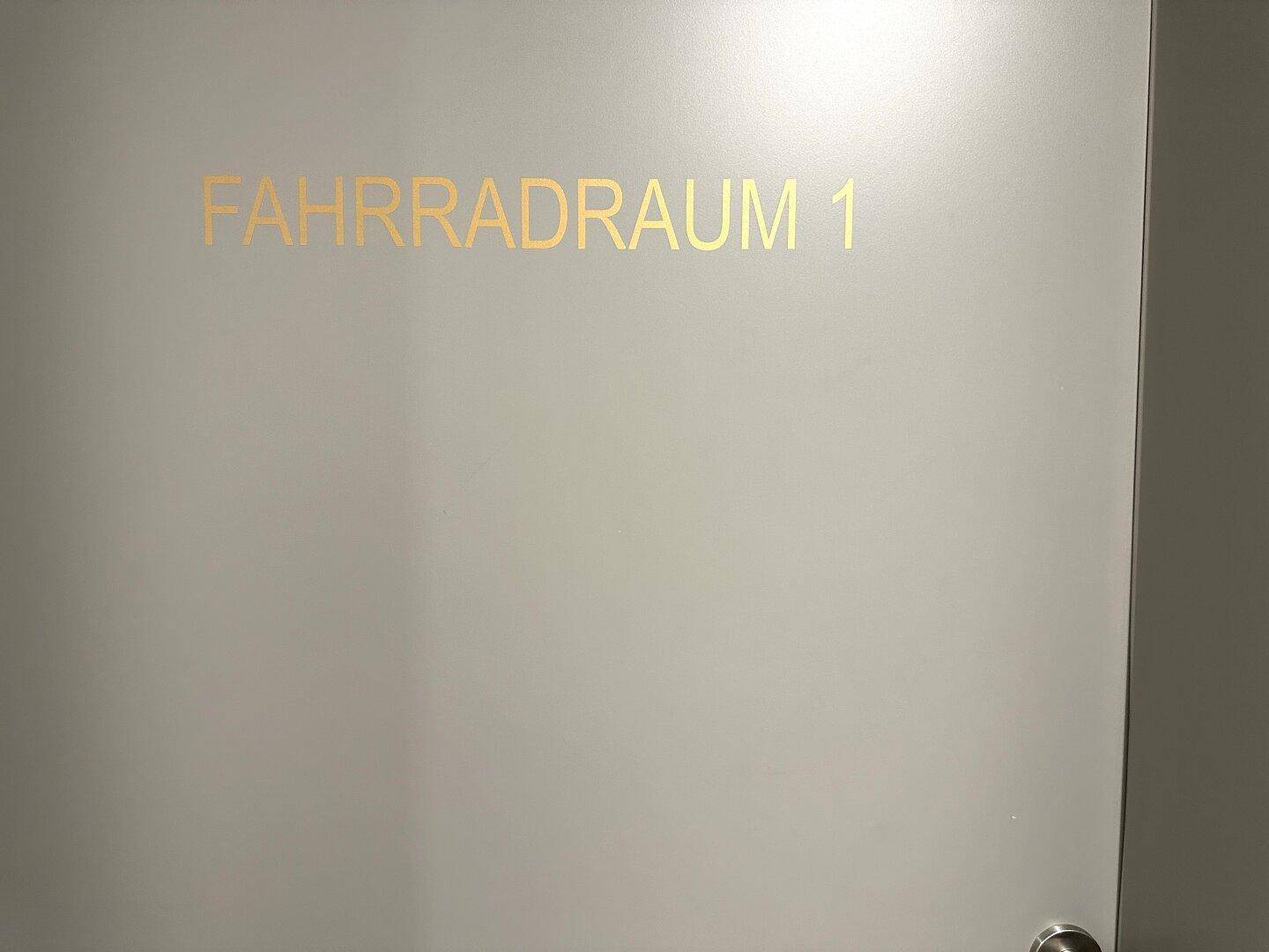 Eine Tür mit der goldenen Aufschrift "Fahrradraum 1" in einem modernen Gebäude.