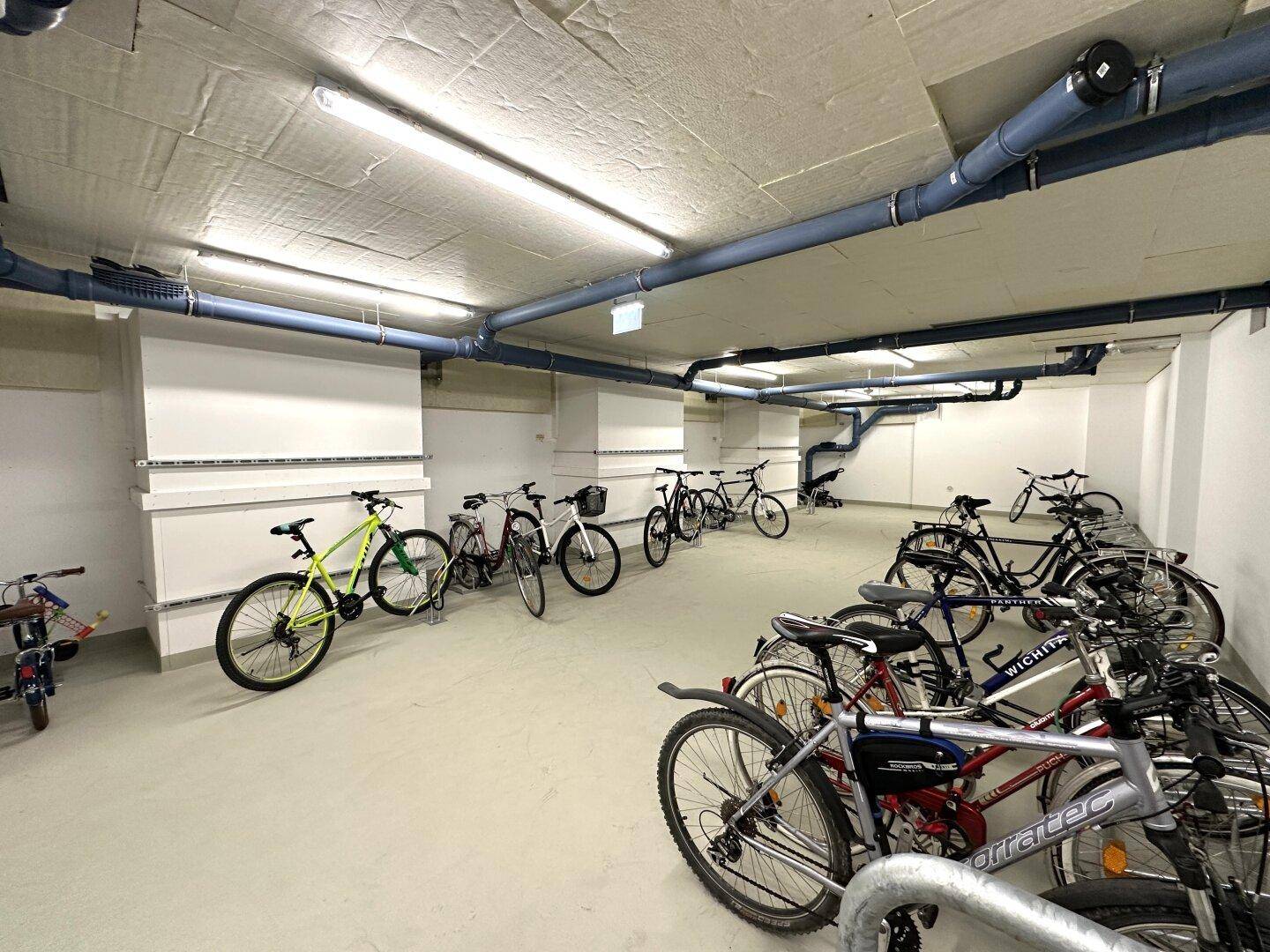 Geräumiger Fahrradraum mit vielen abgestellten Fahrrädern und sichtbaren Rohren an der Decke.