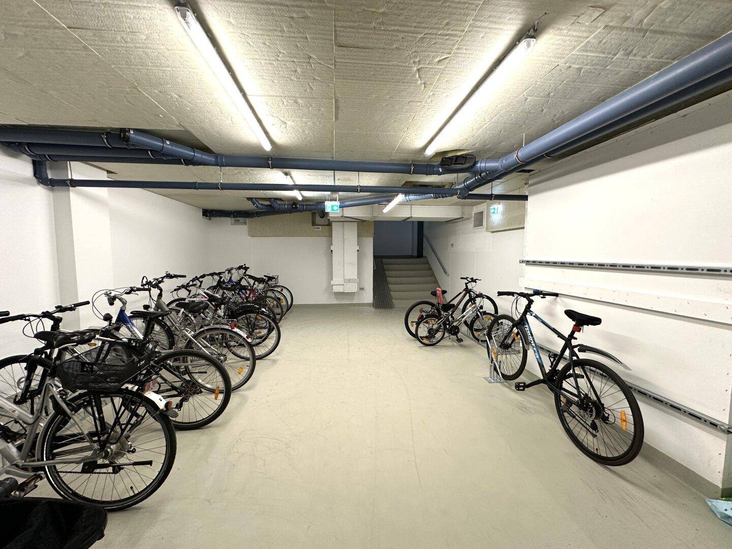 Heller Fahrradraum mit zahlreichen Fahrrädern und funktionaler Beleuchtung in einem modernen Gebäude.