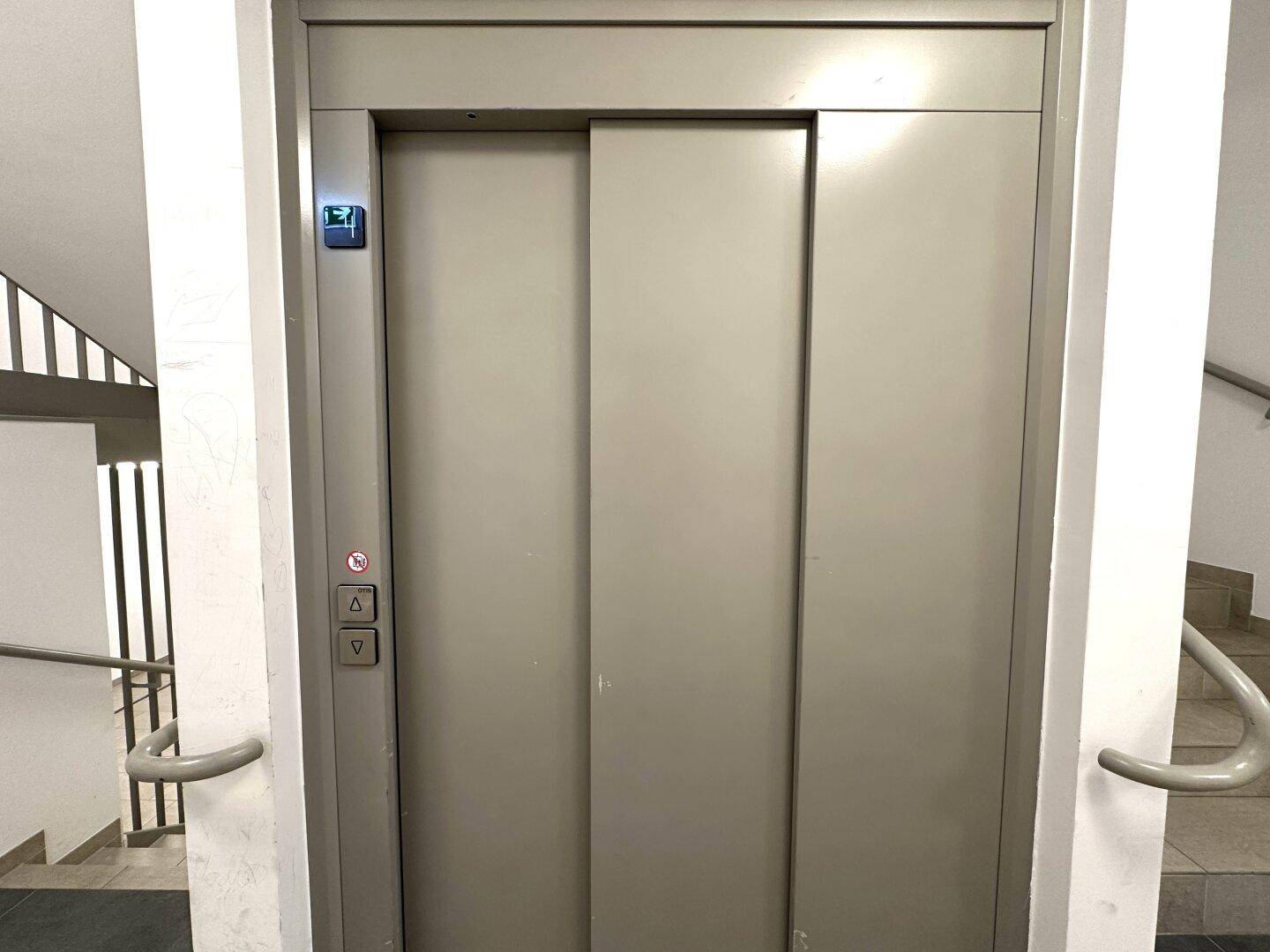 Geschlossene Aufzugstür in einem Treppenhaus mit Handläufen und Fliesenboden.