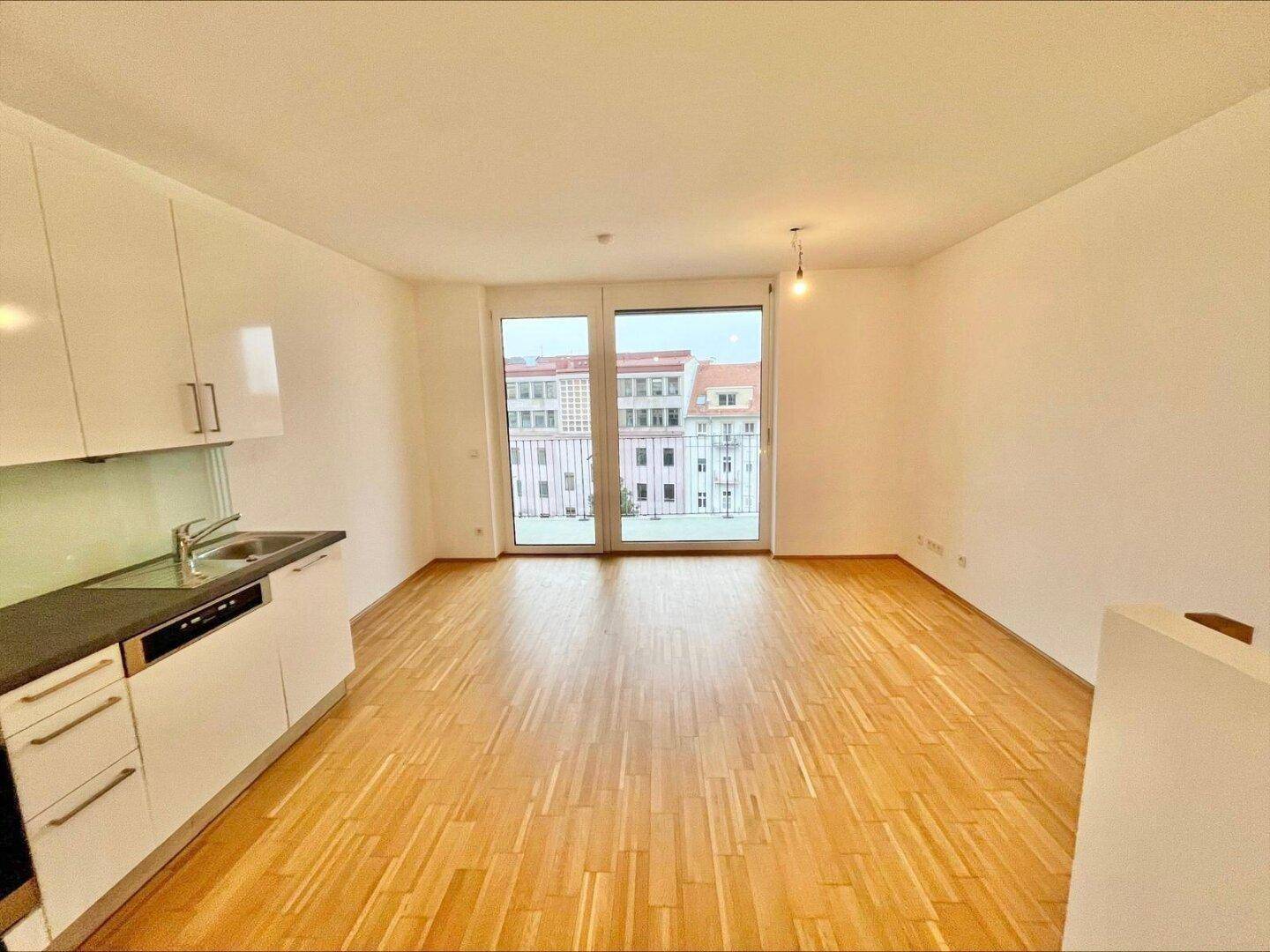 Heller Wohnbereich mit integrierter Küchenzeile, Parkettboden und Zugang zum Balkon.