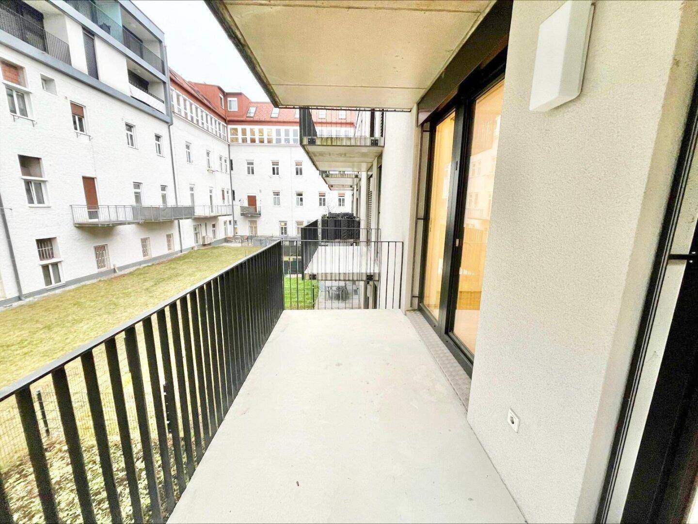Langer Balkon mit schwarzem Metallgeländer und Blick auf eine grüne Innenhofanlage.