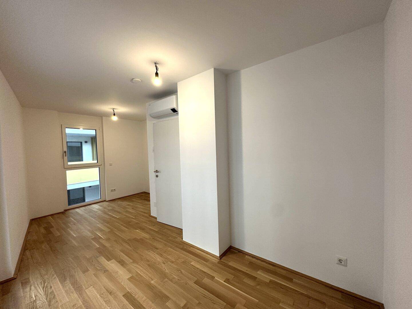 Helles Schlafzimmer mit Holzboden, Klimaanlage und Fenster, das viel Tageslicht hereinlässt.