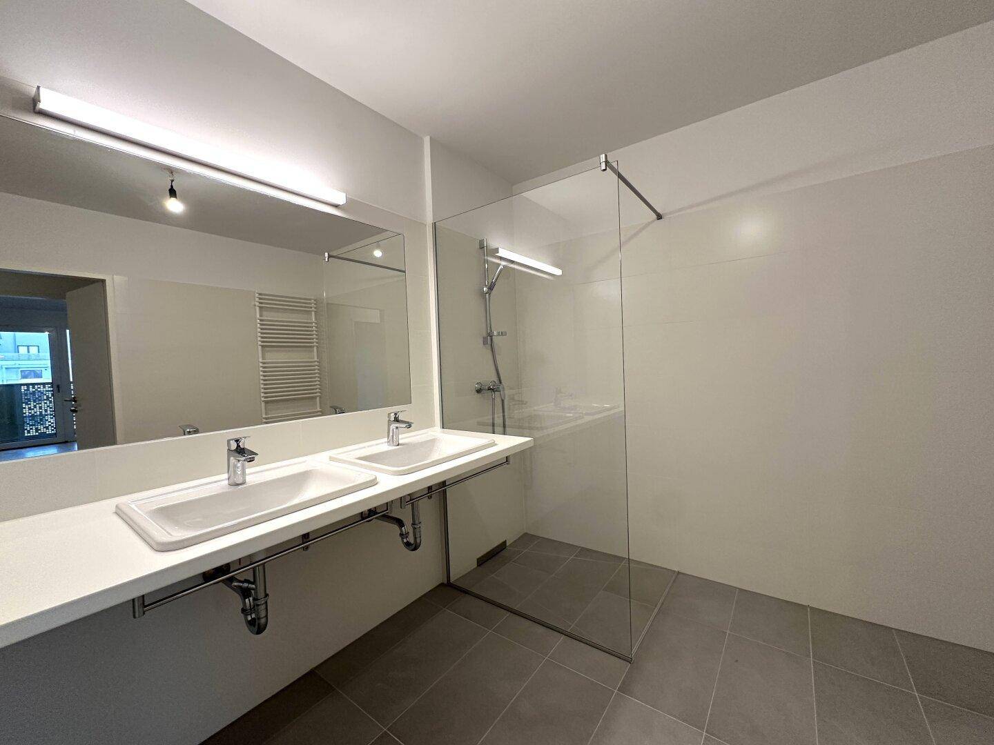 Helles Badezimmer mit zwei Waschbecken, großem Spiegel und geräumiger, moderner Dusche.