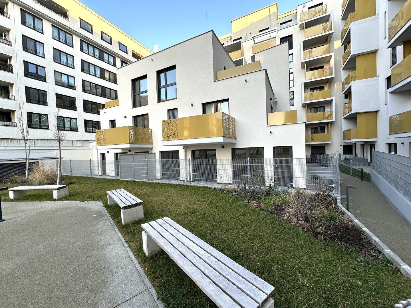Moderne Wohnanlage mit begrüntem Innenhof, Sitzbänken und gelben Balkonakzenten.