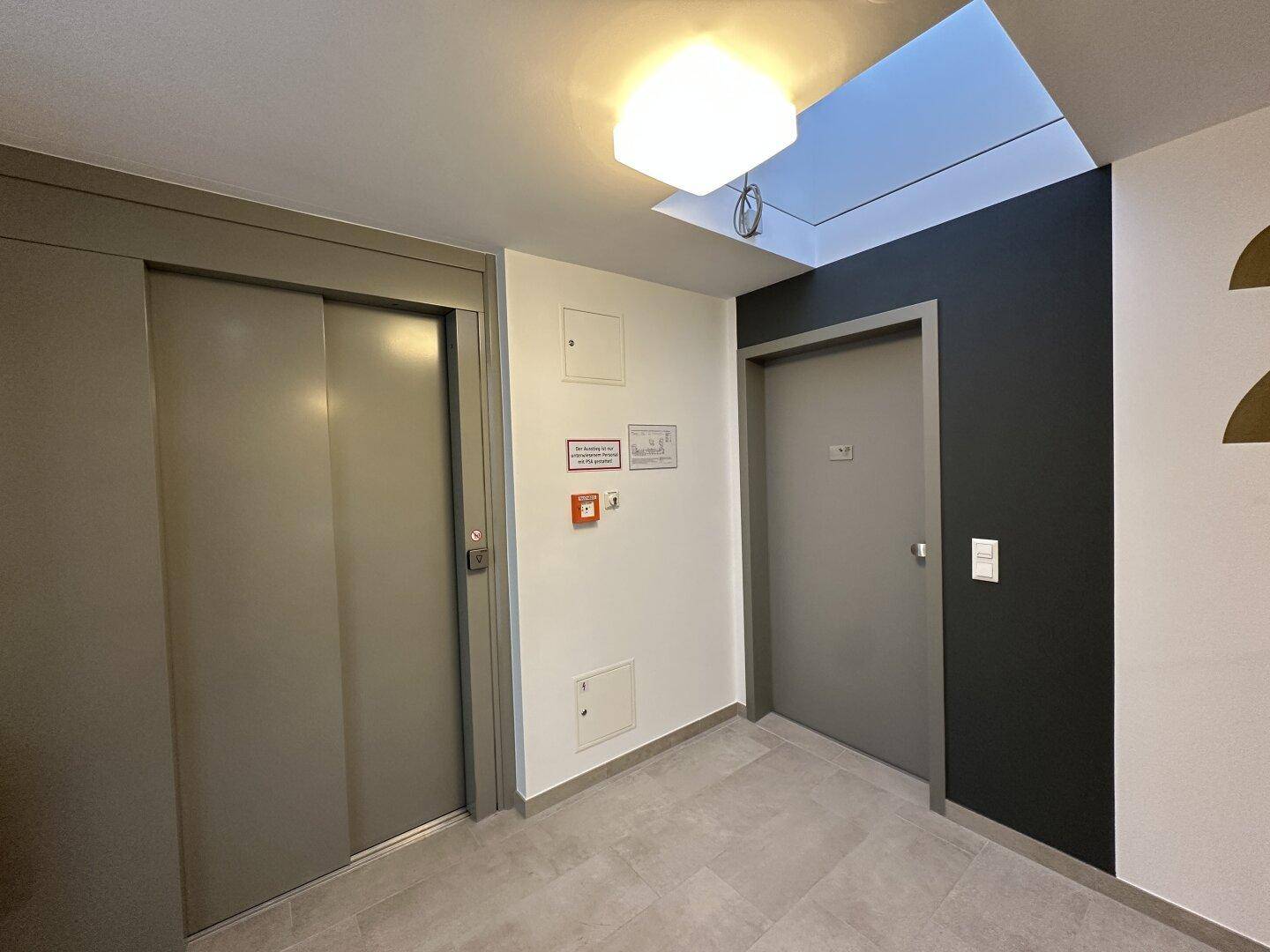 Heller Flur mit Aufzugstür, weiteren Türen und einem Oberlicht für natürliches Licht.