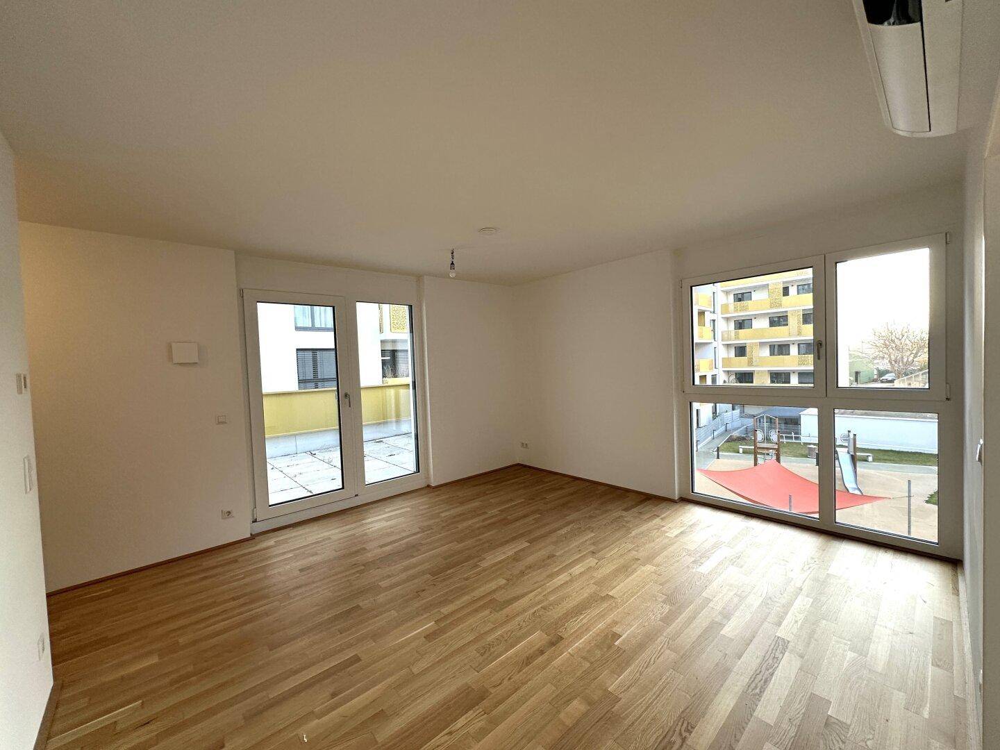 Geräumiges Wohnzimmer mit Holzboden, großen Fenstern und Blick auf einen Spielplatz.