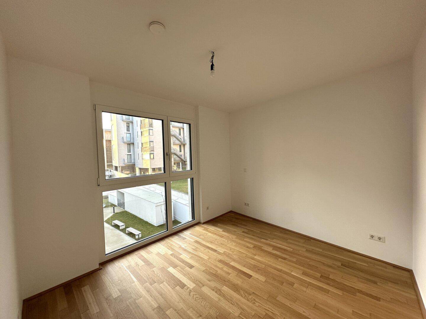 Helles Schlafzimmer mit Holzboden und großem Fenster, das viel Tageslicht hereinlässt.