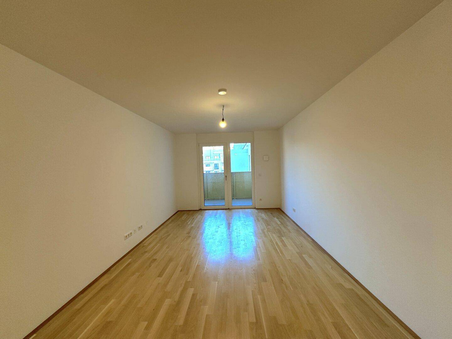 Geräumiger Wohnbereich mit Holzboden und großen Fenstern zum Balkon.