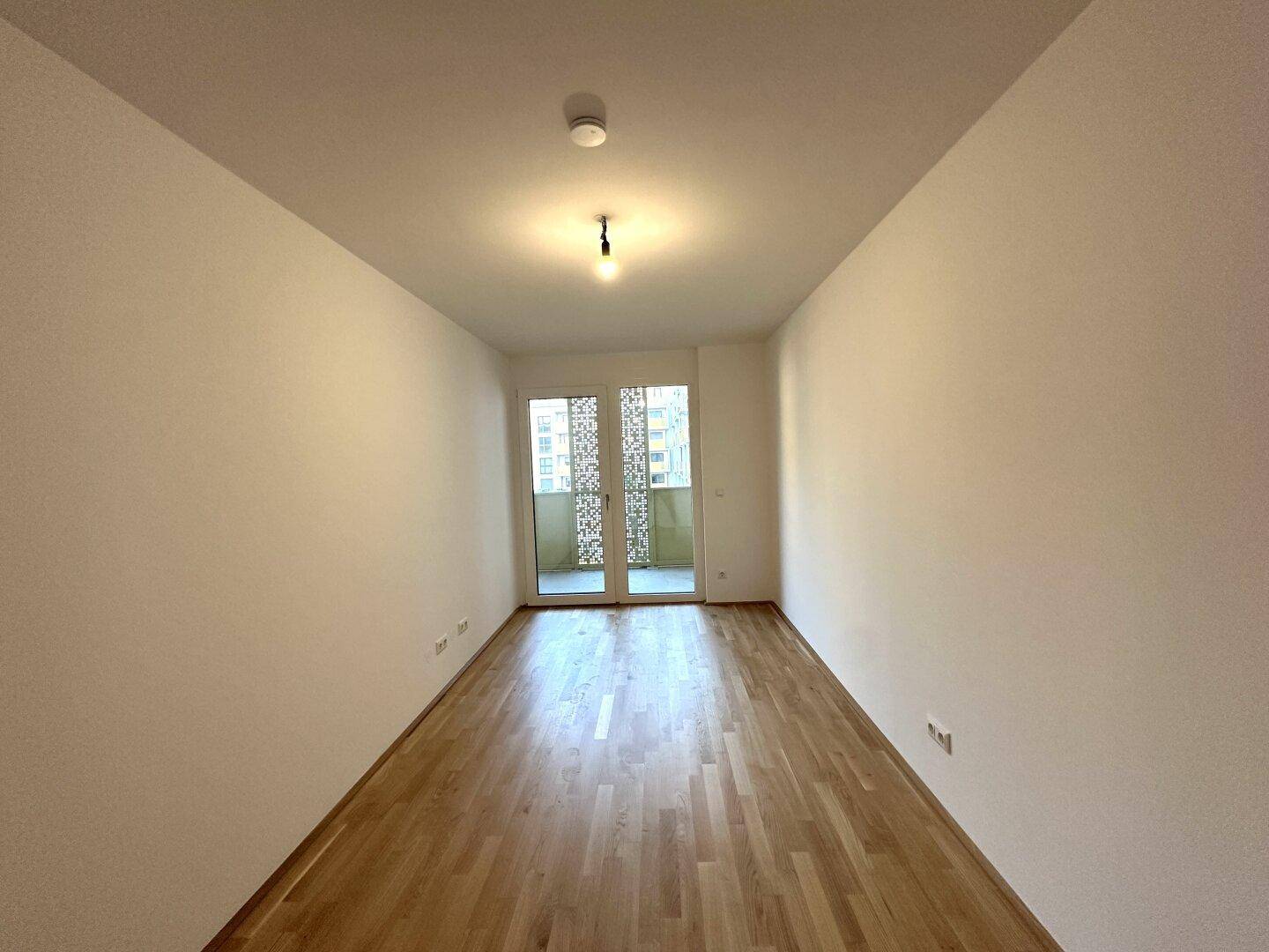 Langes, helles Zimmer mit Parkettboden und Zugang zu einem Balkon.