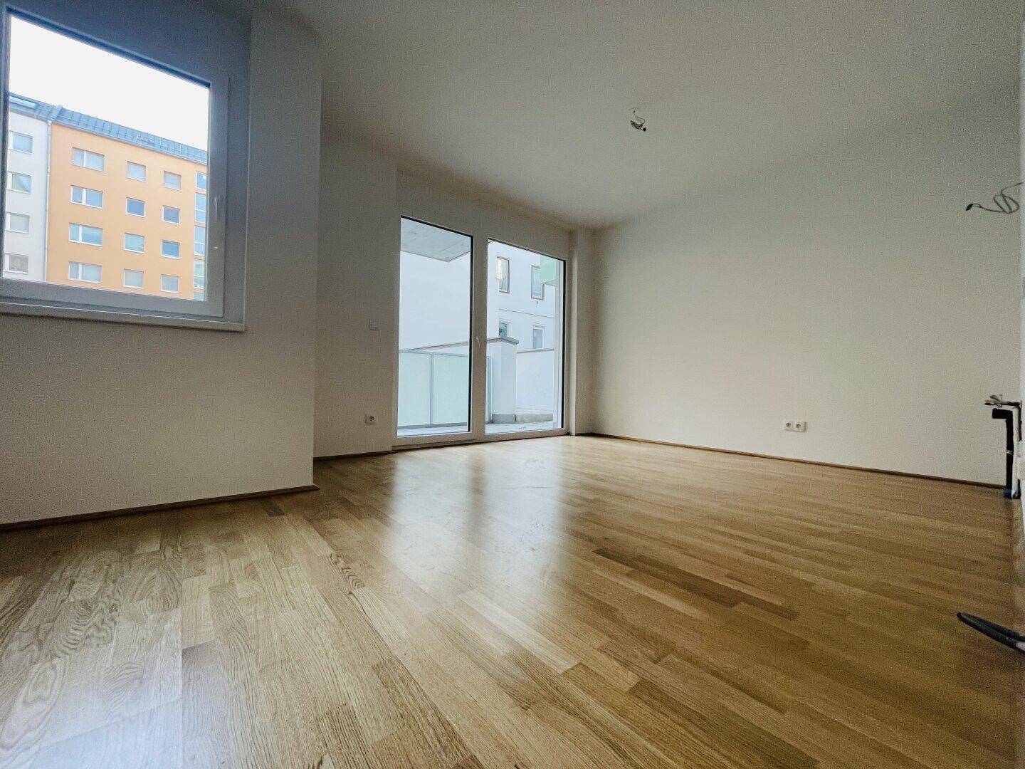 Helles Zimmer mit Parkettboden, weißen Wänden und Zugang zu einem Balkon.