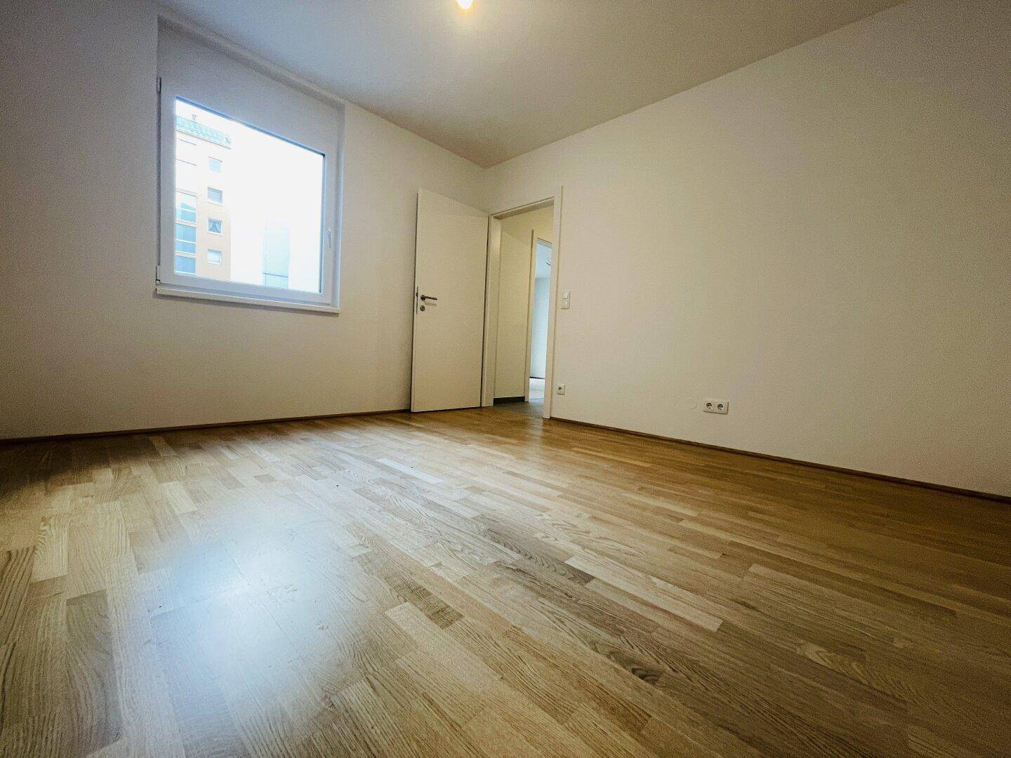Leeres Schlafzimmer mit hellem Parkettboden, weißen Wänden und einem großen Fenster.