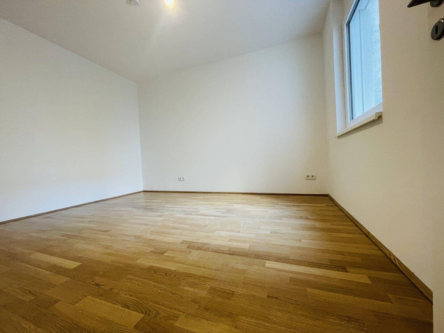 Helles Schlafzimmer mit Parkettboden, weißen Wänden und einem Fenster, das viel Licht hereinlässt.