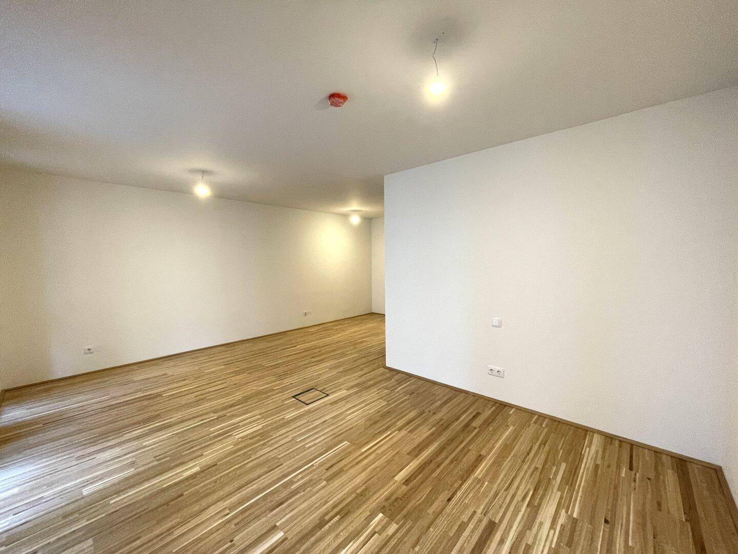 Geräumiges, leeres Wohnzimmer mit hellem Parkettboden und weißen Wänden.