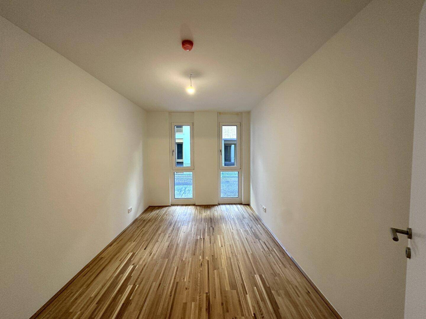 Helles Zimmer mit Parkettboden und zwei hohen Fenstern, die den Raum mit natürlichem Licht füllen.