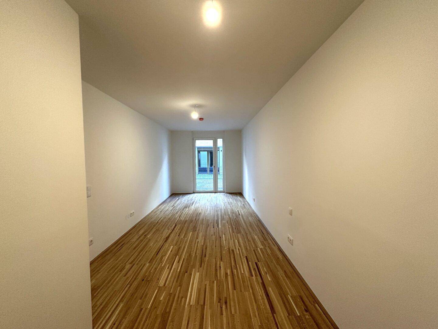 Langes, helles Zimmer mit Parkettboden und Fenstertür, die den Raum mit natürlichem Licht versorgt.