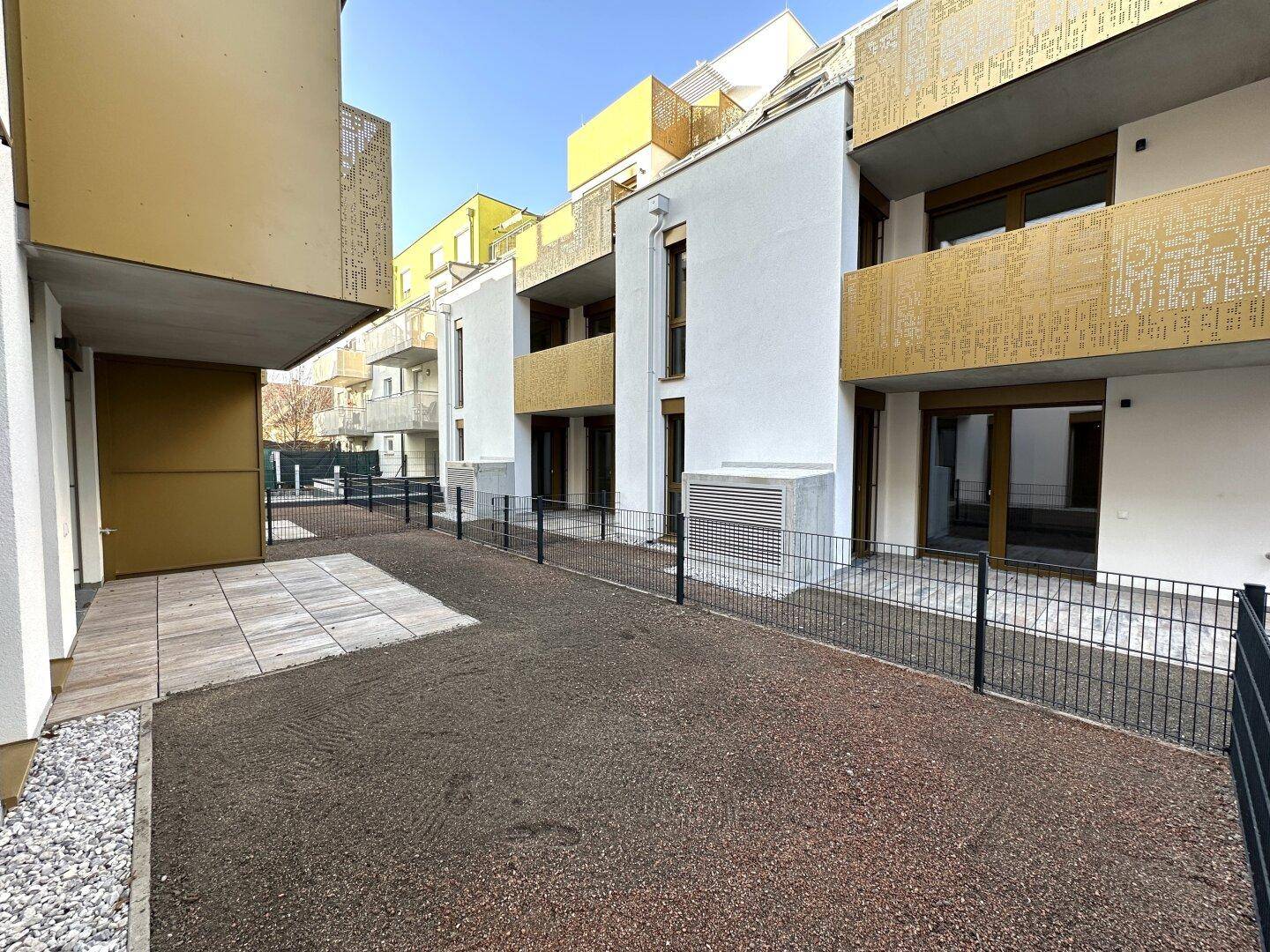 Außenansicht des Neubaus mit privaten Gartenbereichen und modernen Balkonen in goldener Farbe.