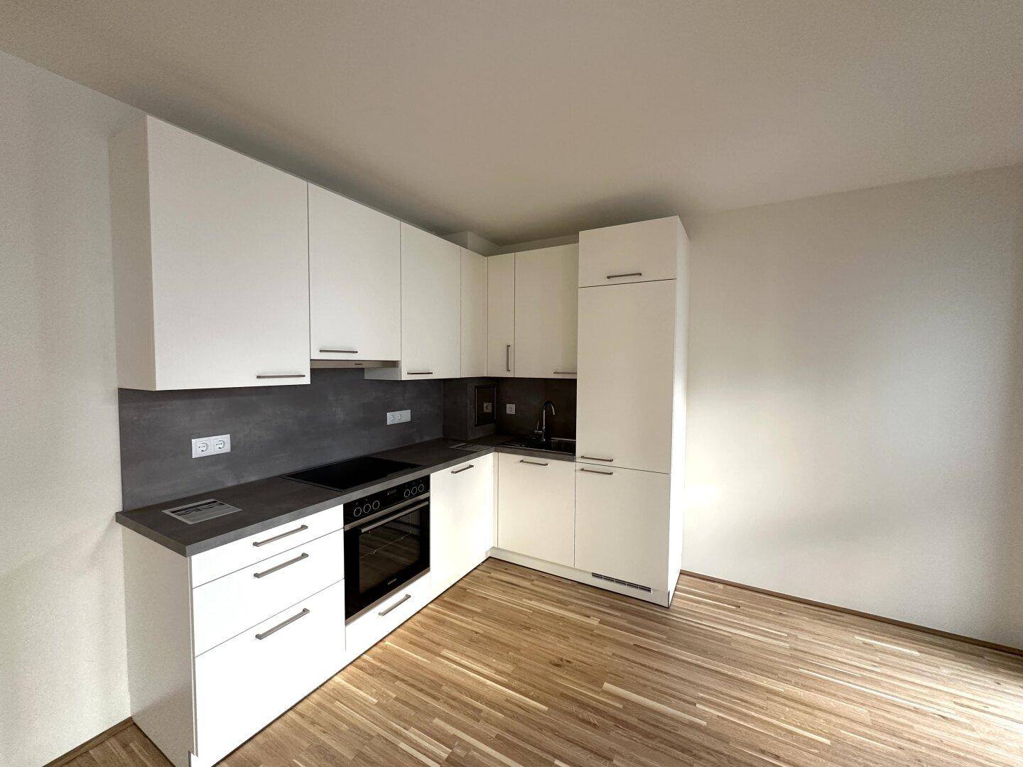 Moderne Einbauküche in L-Form mit weißen Schränken, grauer Arbeitsplatte und integrierten Elektrogeräten.