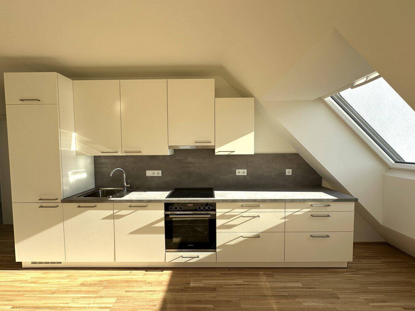 Moderne Einbauküche mit weißen Schränken, dunkler Arbeitsplatte und integrierten Geräten unter Dachschräge.